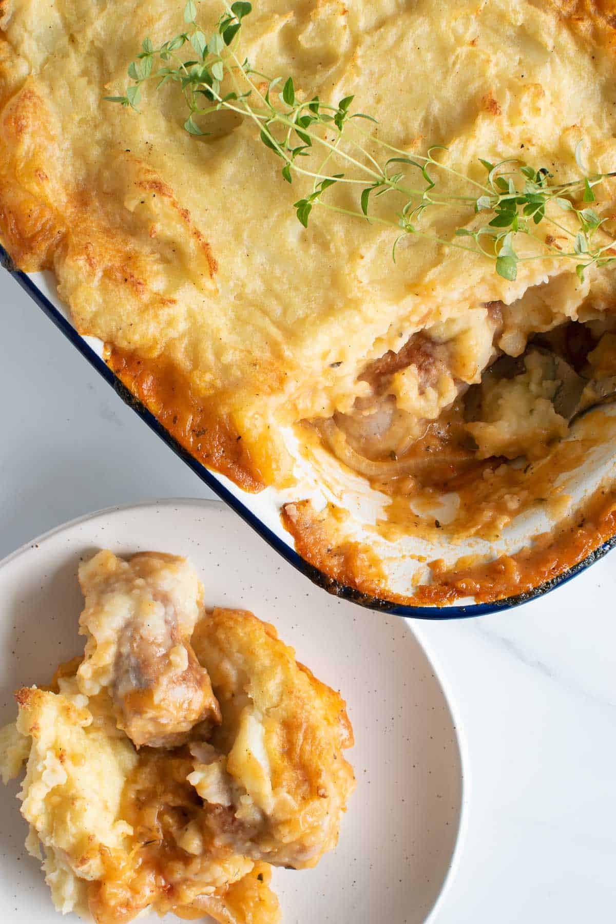 A pie dish filled with sausage and mashed potato pie, with a serving plated up on the side.