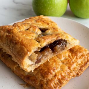 Two puff pastry hand pies with apple filling, with apples in the background.