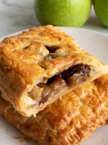 Two puff pastry hand pies with apple filling, with apples in the background.