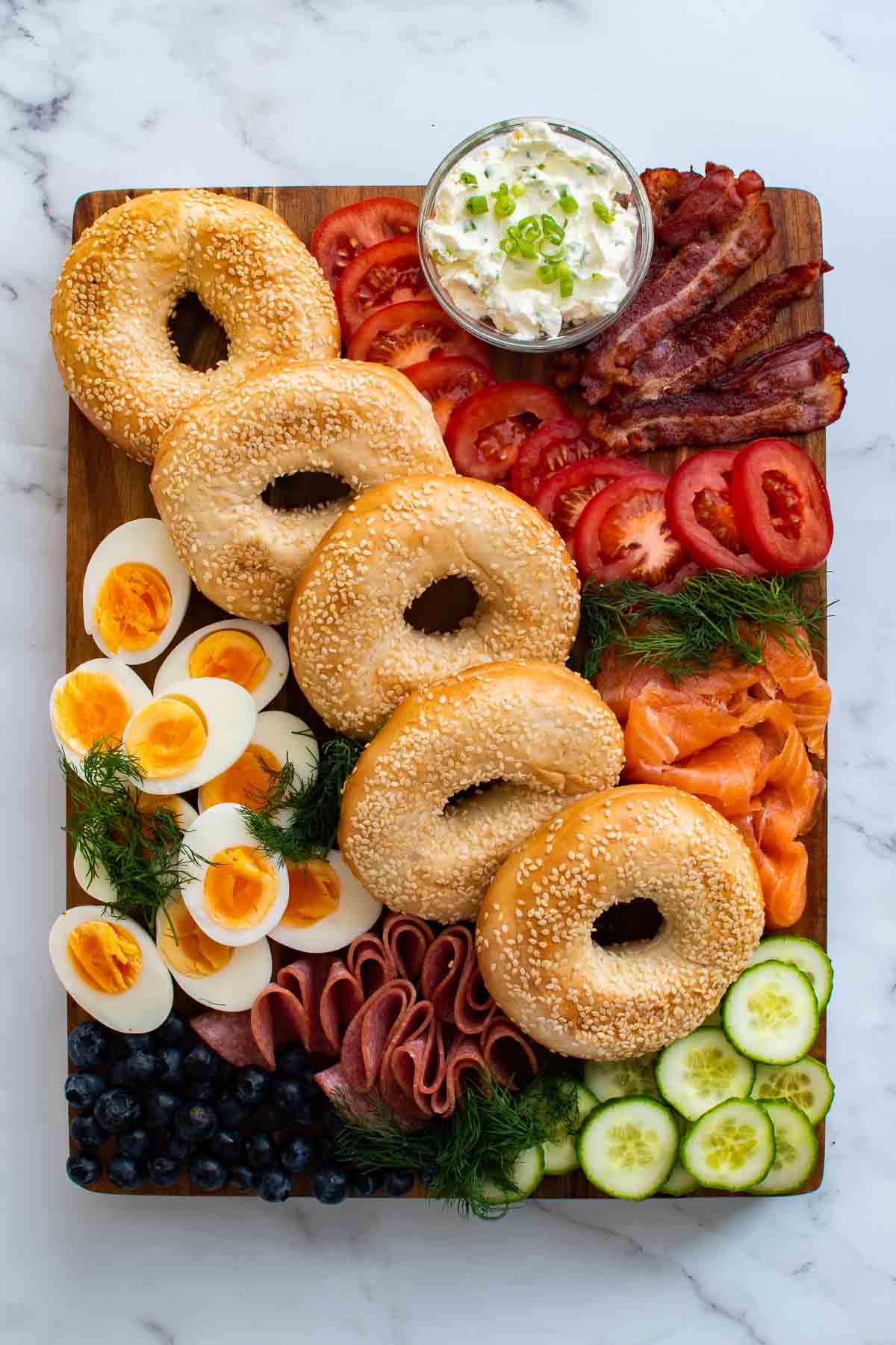 A bagel board with cream cheese spread, bacon, tomatoes, cucumber, smoked salmon, boiled eggs, salami, dill and blueberries,