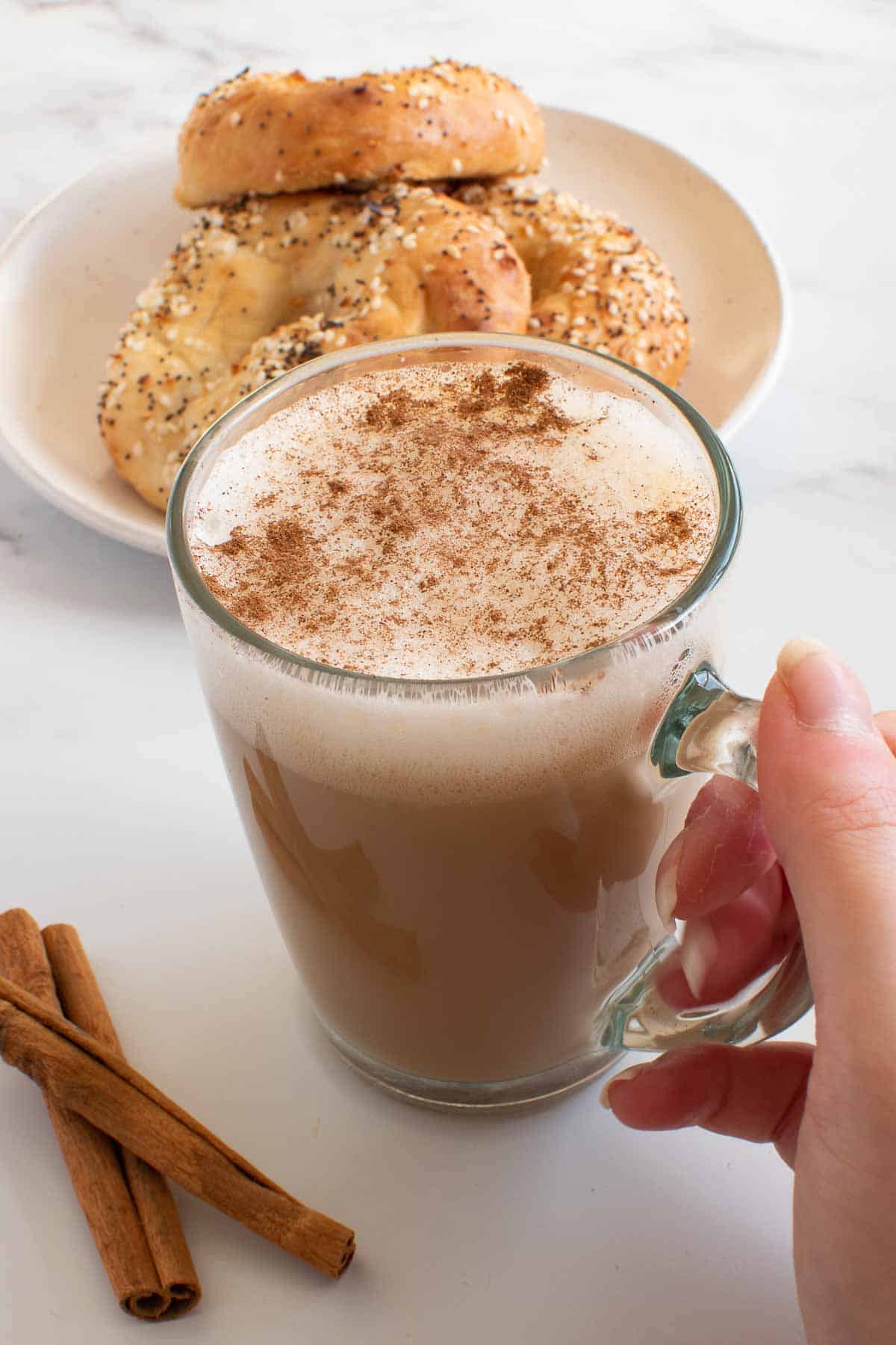 A hand picking up a cup of eggnog latte, with bagels in the background.