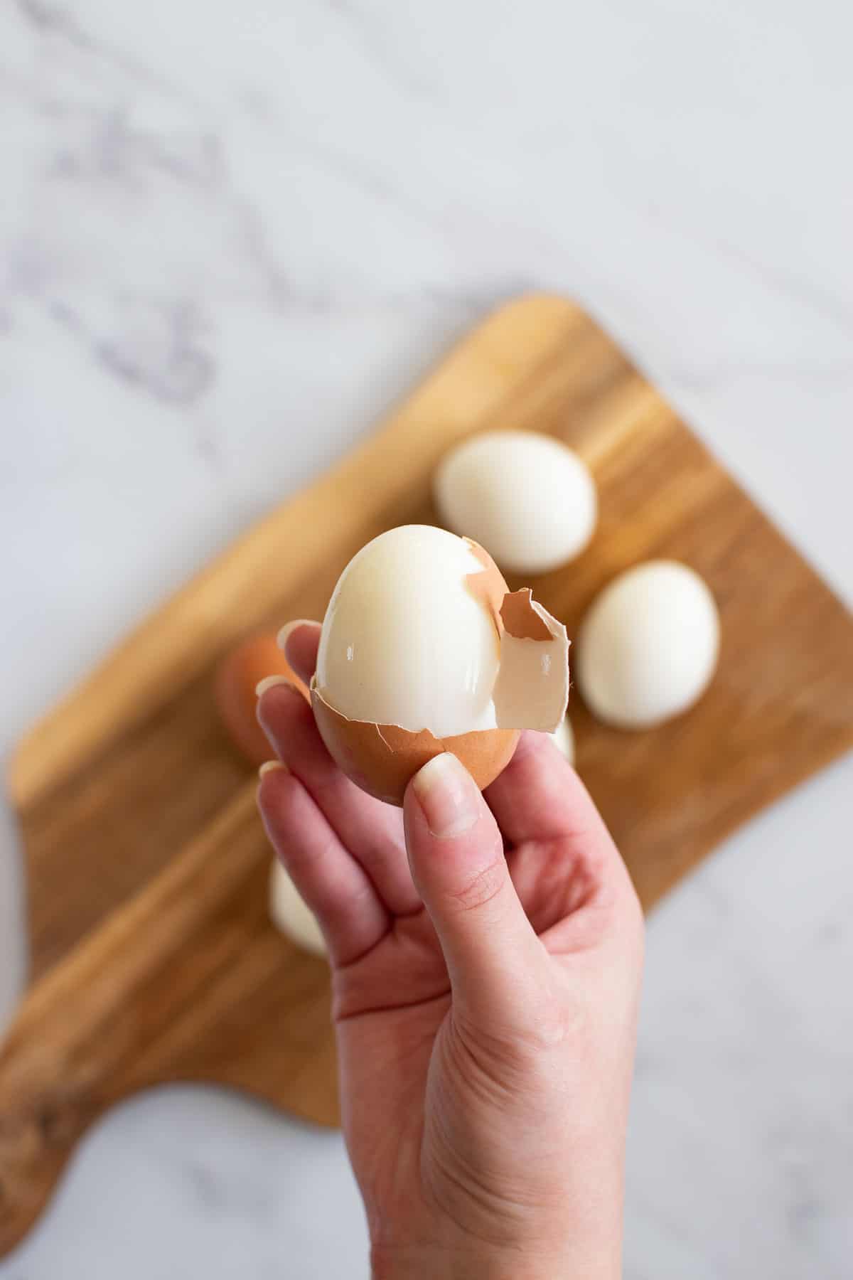 A hand holding up a half peeled hard boiled egg, with more eggs in the background.