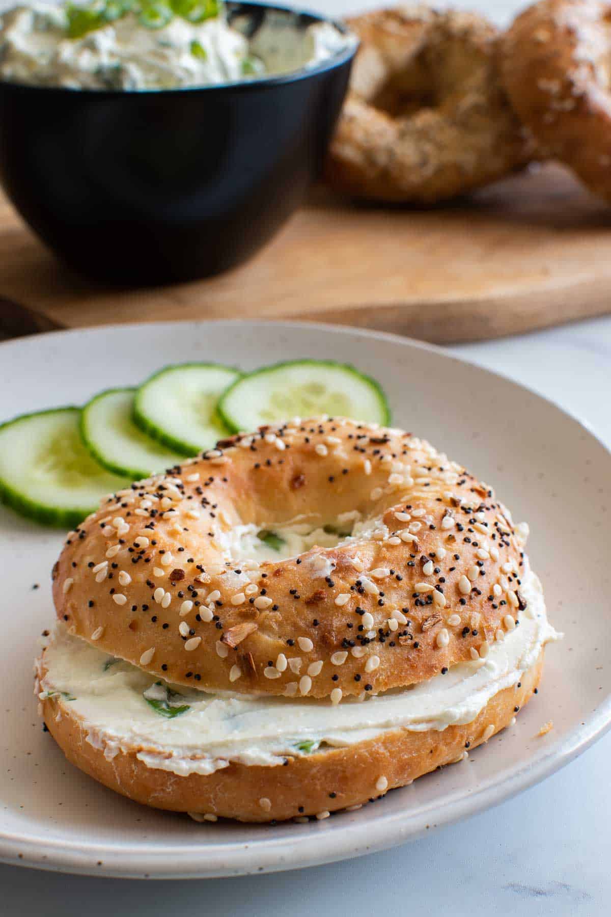 A bagel with cream cheese and scallions, with cucumbers and bagels in the background.