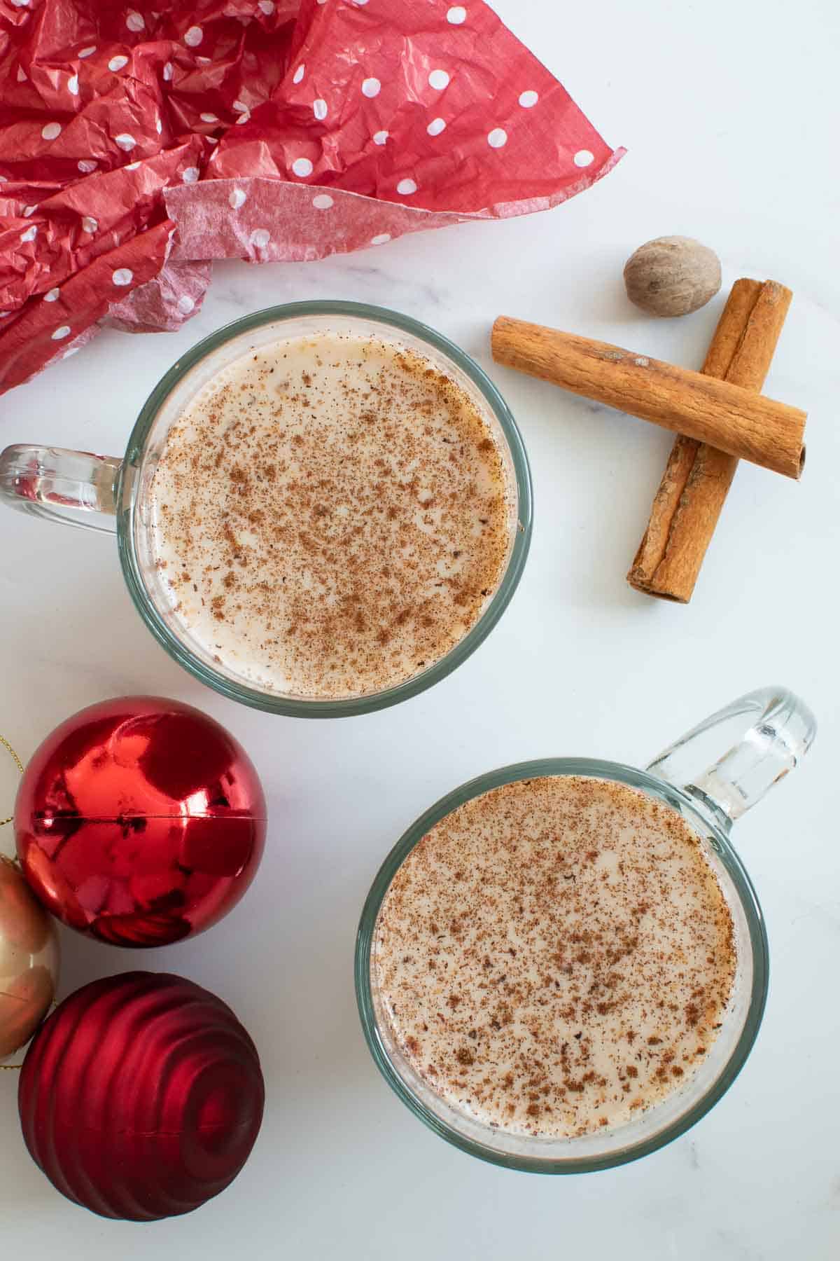 Two cups of eggnog, with Christmas tree decorations and cinnamon sticks on the side.