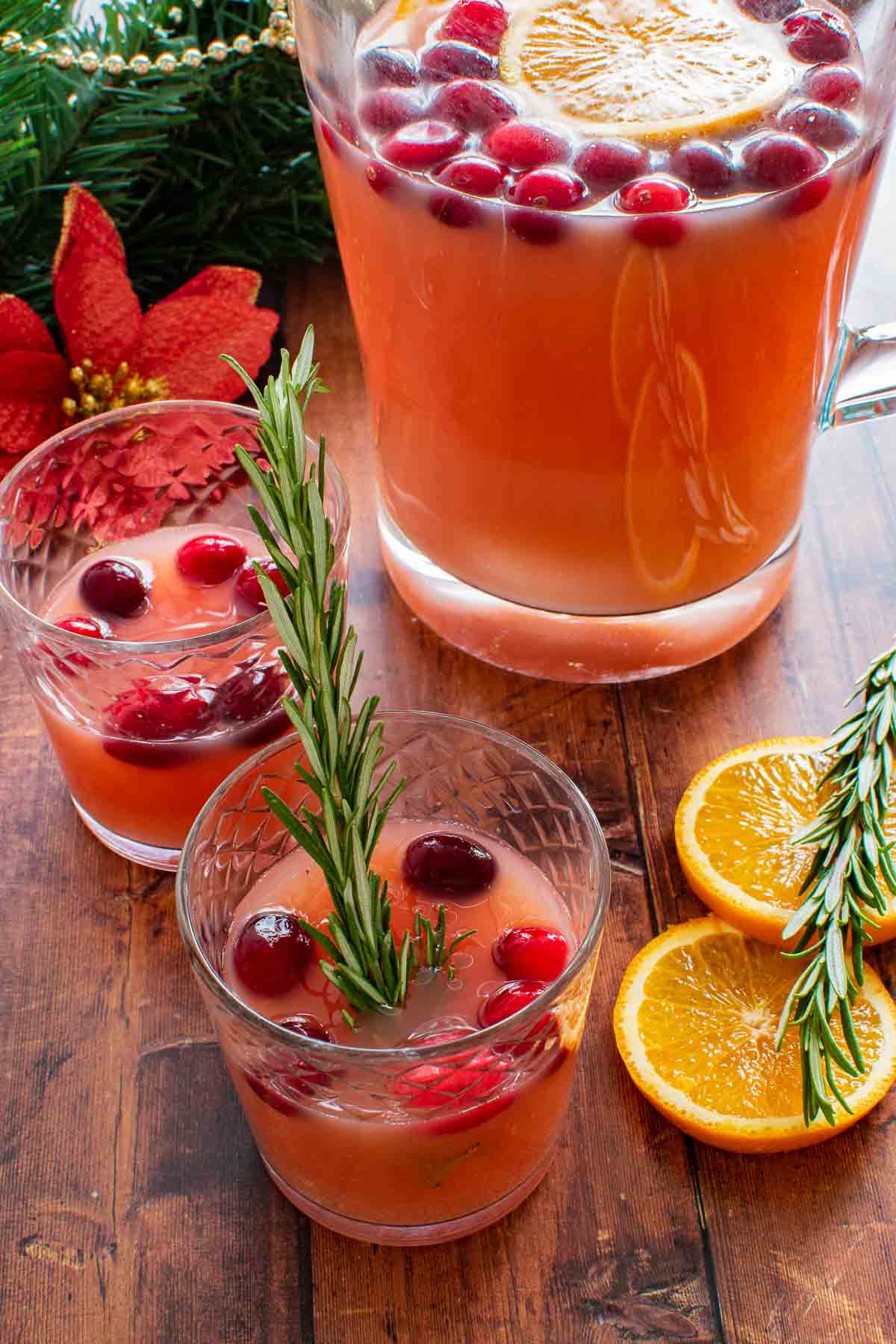 A pitcher of punch with two glasses on the side. Garnished with orange slices and cranberries.