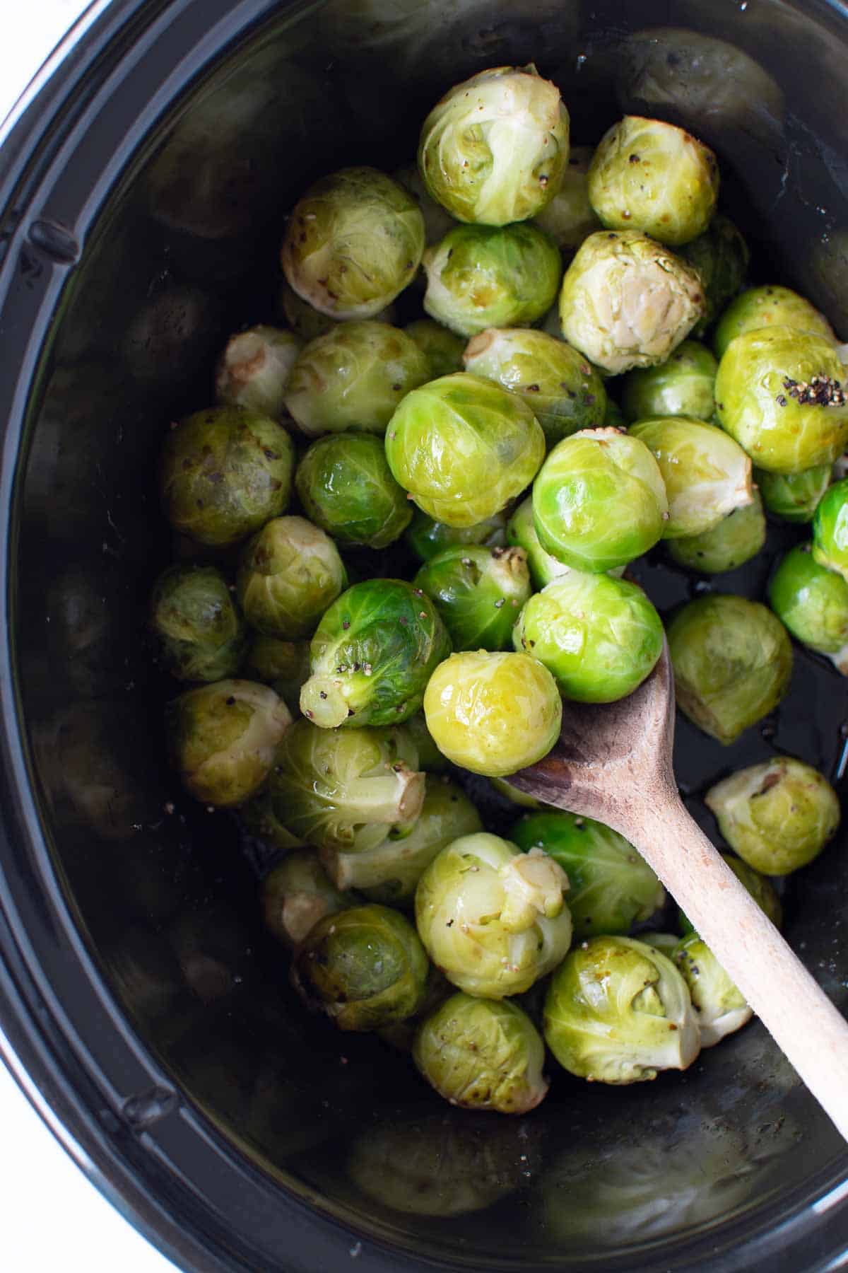 Brussels sprouts in a slow cooker. A wooden spoon is lifting some of them up.