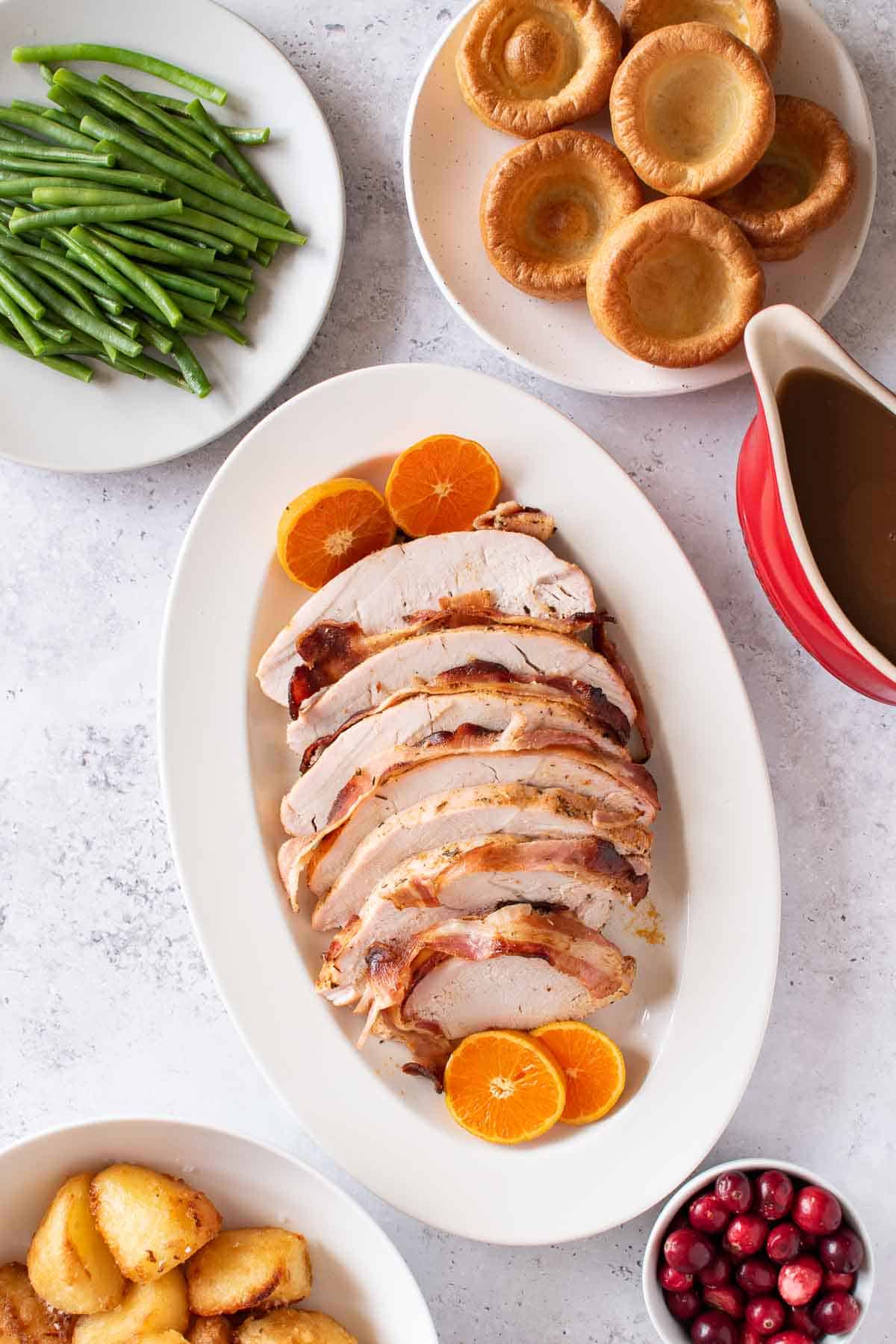 Sliced turkey breast on a serving platter, with yorkshire puddings, gravy, green beans, cranberries and potatoes on the side.