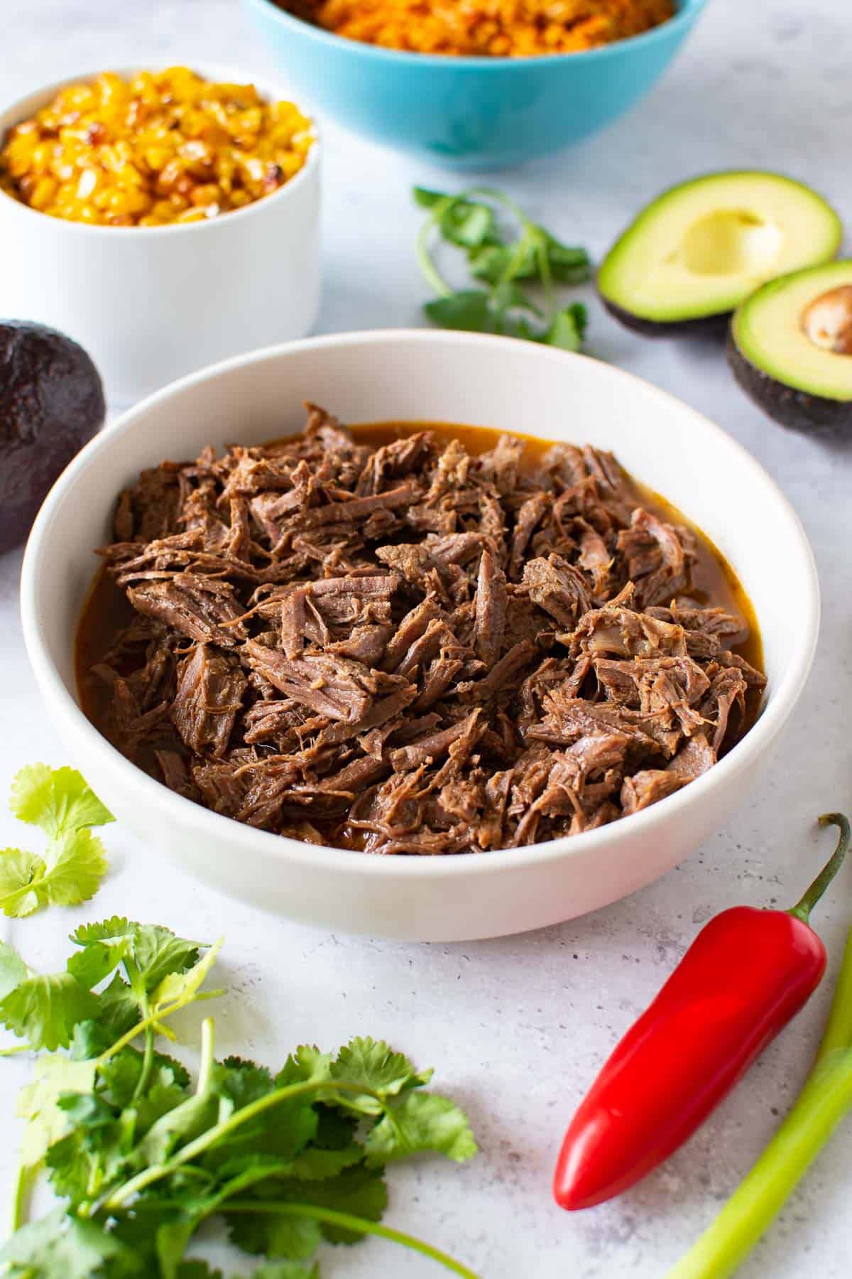 Mexican shredded beef in a bowl, with vegetables and side dishes around.