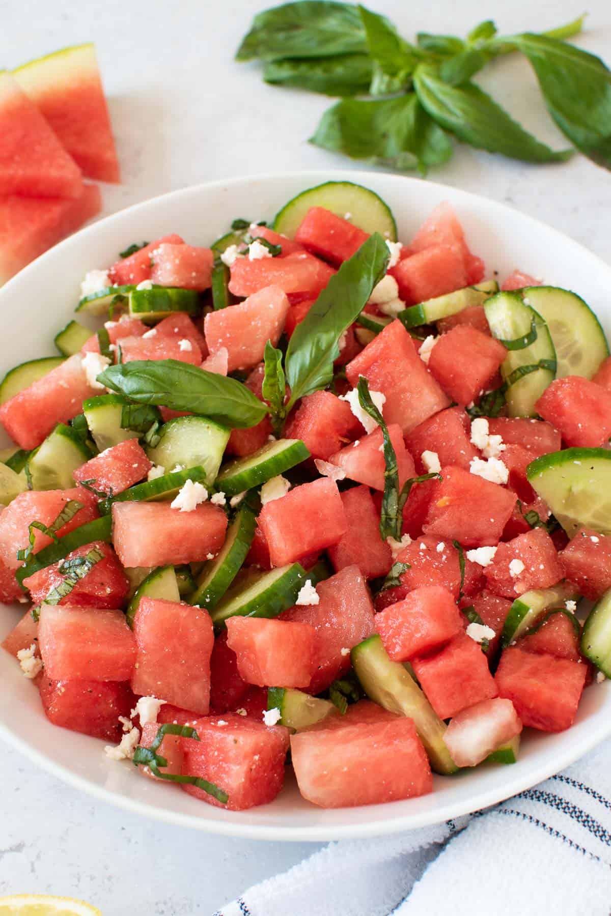 Cucumber and watermelon salad with feta cheese in a salad bowl, with basil and melon in the background.