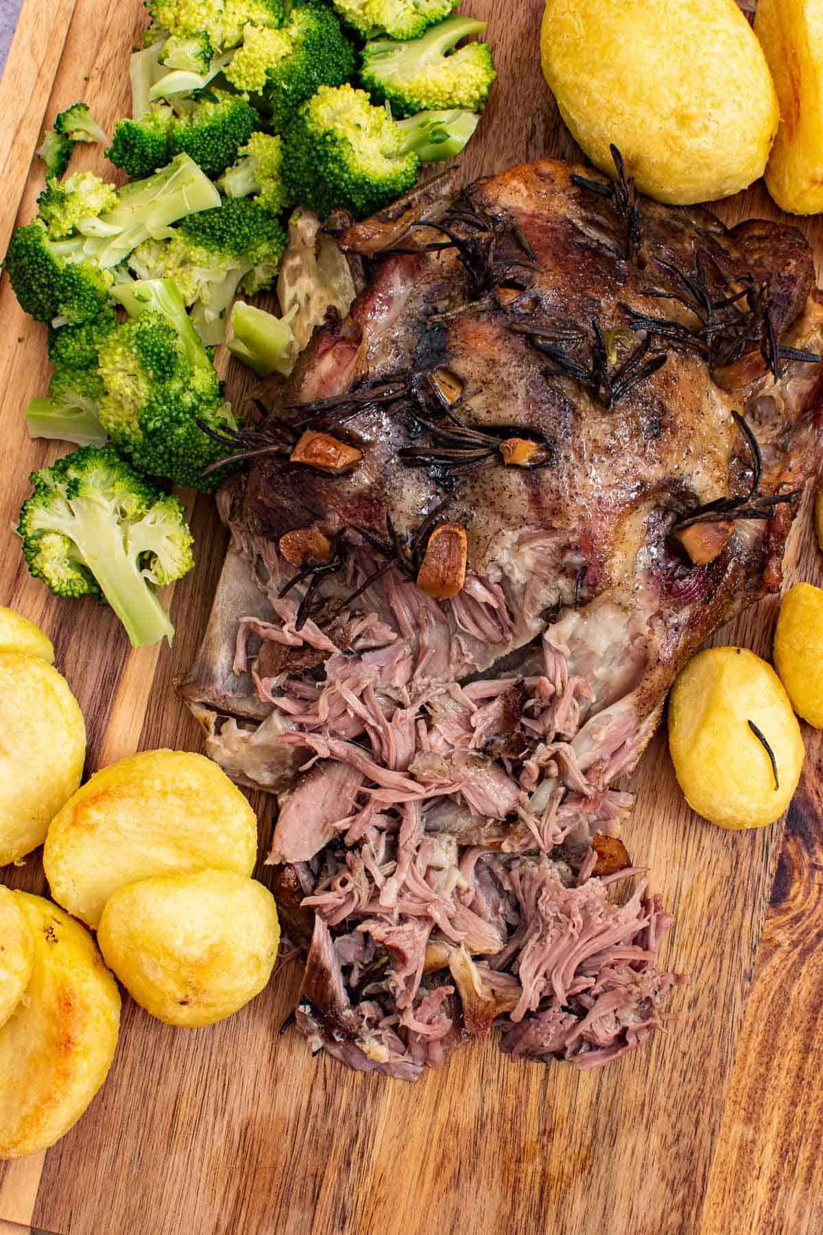 Slow cooked lamb shoulder on a wooden board with broccoli and potatoes on the side.