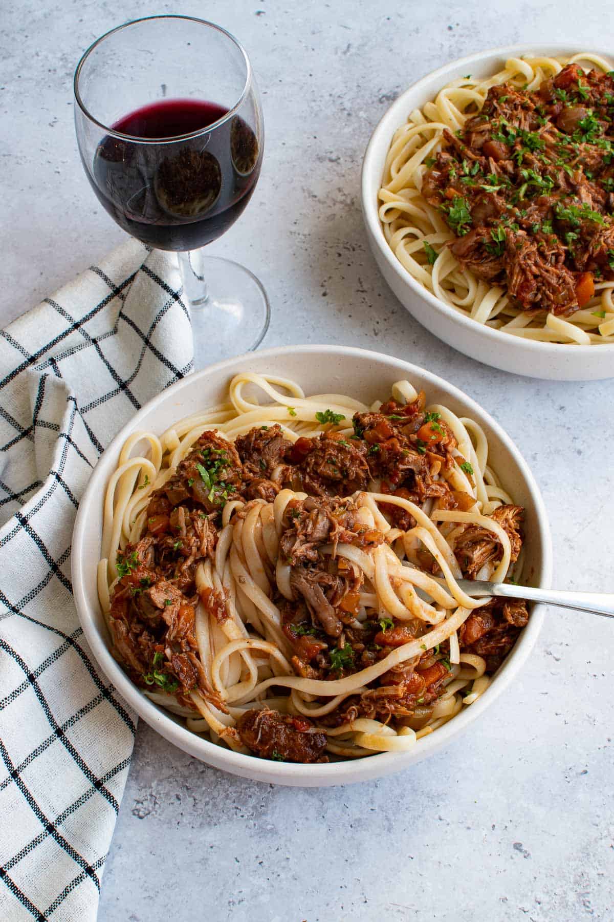 Two bowls of pasta with lamb ragu on a table, with a glass of red wine on the side.