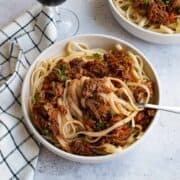 A close up of a bowl of linguine wih slow cooked lamb ragu. A dish cloth and a glass of wine is on the side.