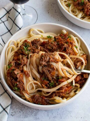 A close up of a bowl of linguine wih slow cooked lamb ragu. A dish cloth and a glass of wine is on the side.